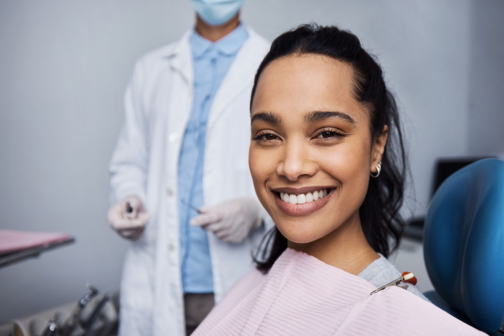 The image shows a woman sitting in a dental chair, wearing a dental bib, with a dentist standing in the background. The dentist is wearing a white coat, a blue shirt, gloves, and a face mask. The setting appears to be a dental clinic.